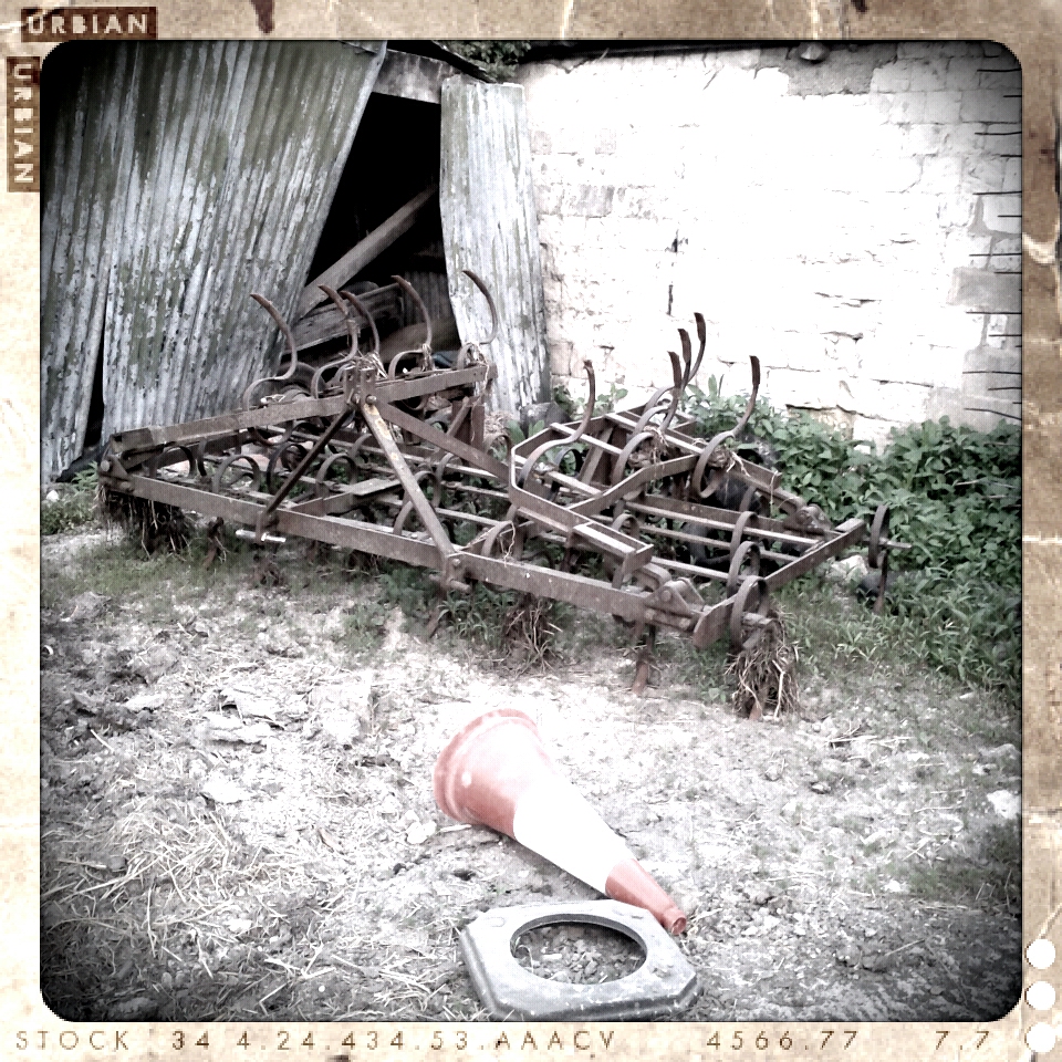 A broken traffic cone lies in front of rusting farm machinery, next to a corrugated barn door in a farmyard