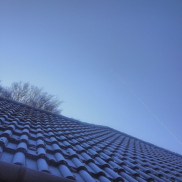 Roof tiles, crsuted with frost, against a bright blue wintry sky with contrails and the outline of a bare tree.