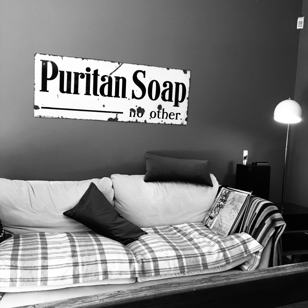 Black and white photo of a sofa underneath an old enamel advertising sign for Puritan Soap