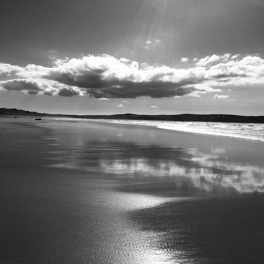 A cloud hovers over an empty beach in bright sunlight, its reflection caught in the wet, shining sand. (Black and white image). 
