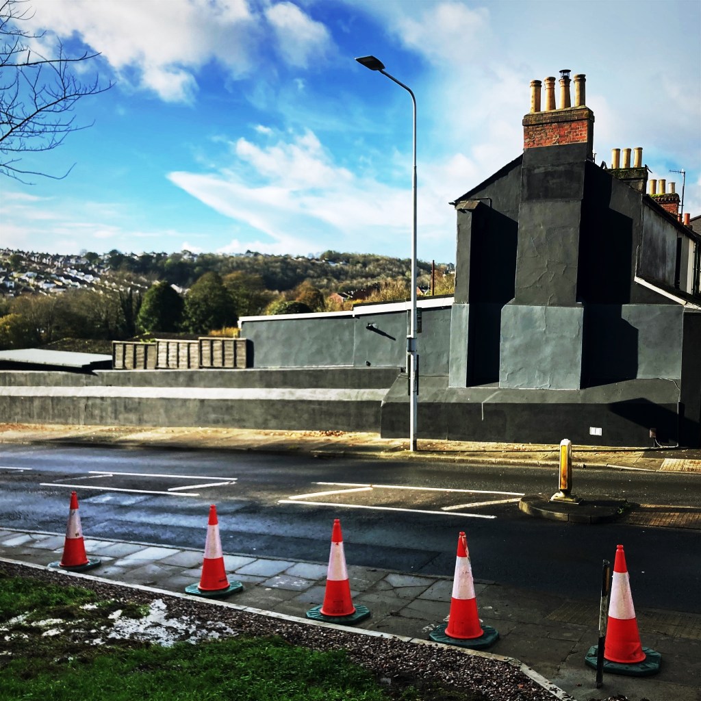Five traffic cones on a pavement next to a main road and Victorian house-end, painted black