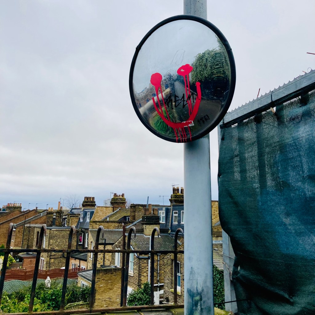 Graffito of a smiley face in dripping pink paint on a visibility mirror on a footbridge across a railway