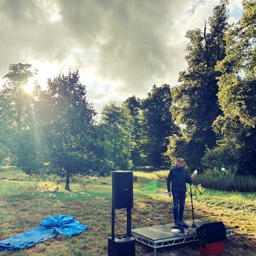 Anthony Wilson stands on a low, open-air stage, framed by sunlight appearing through clouds and trees