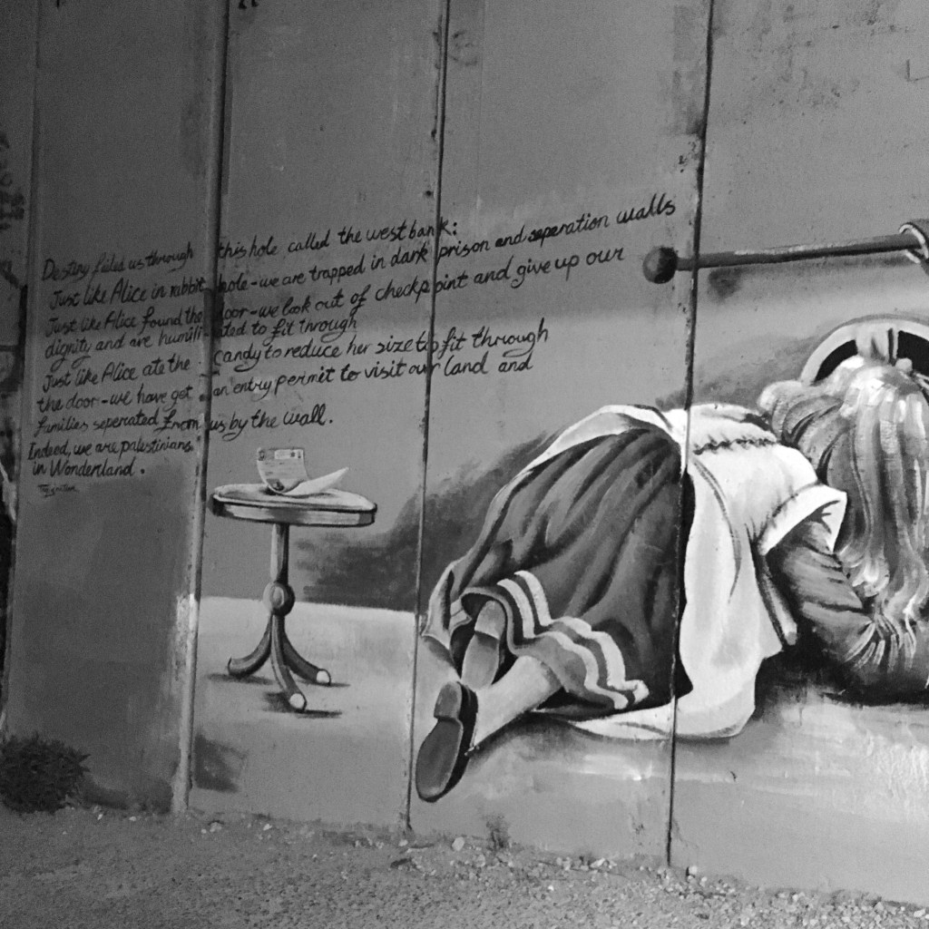 A black and white image of graffiti on the Israeli defence wall depicting Alice in Wonderland attempting to burrow beneath it.