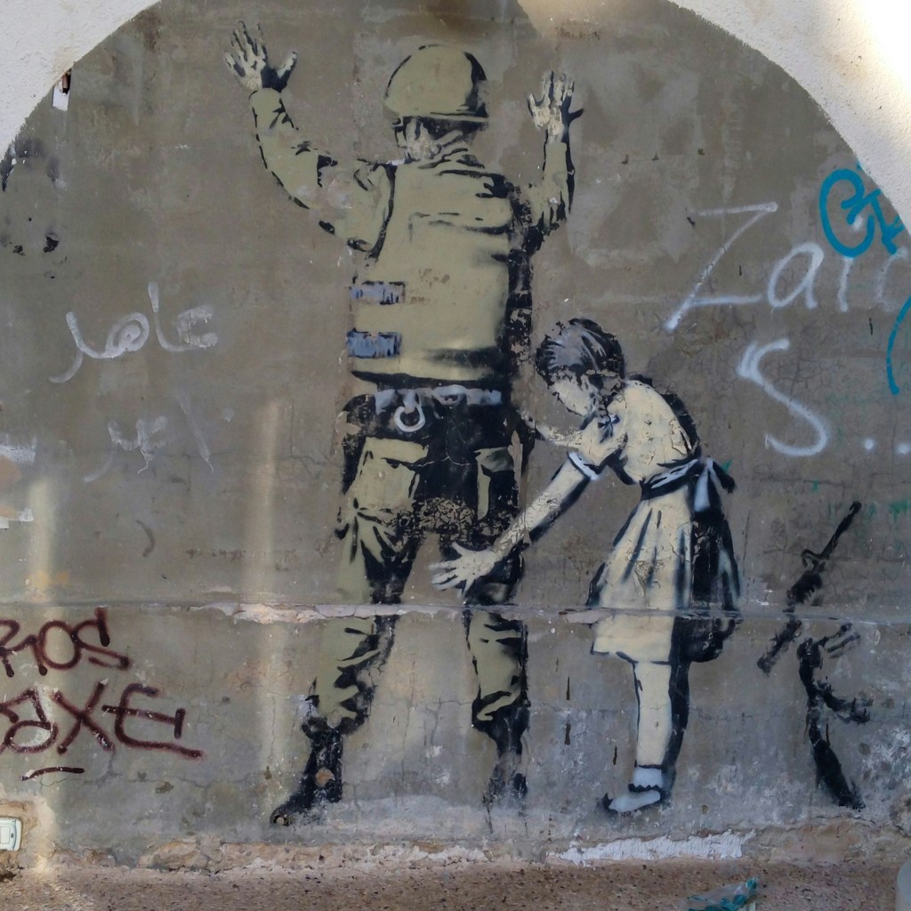 Banksy graffiti on the Israeli defence wall depicting a young girl frisking a soldier from the IDF, his arms raised and hands spread on the wall.