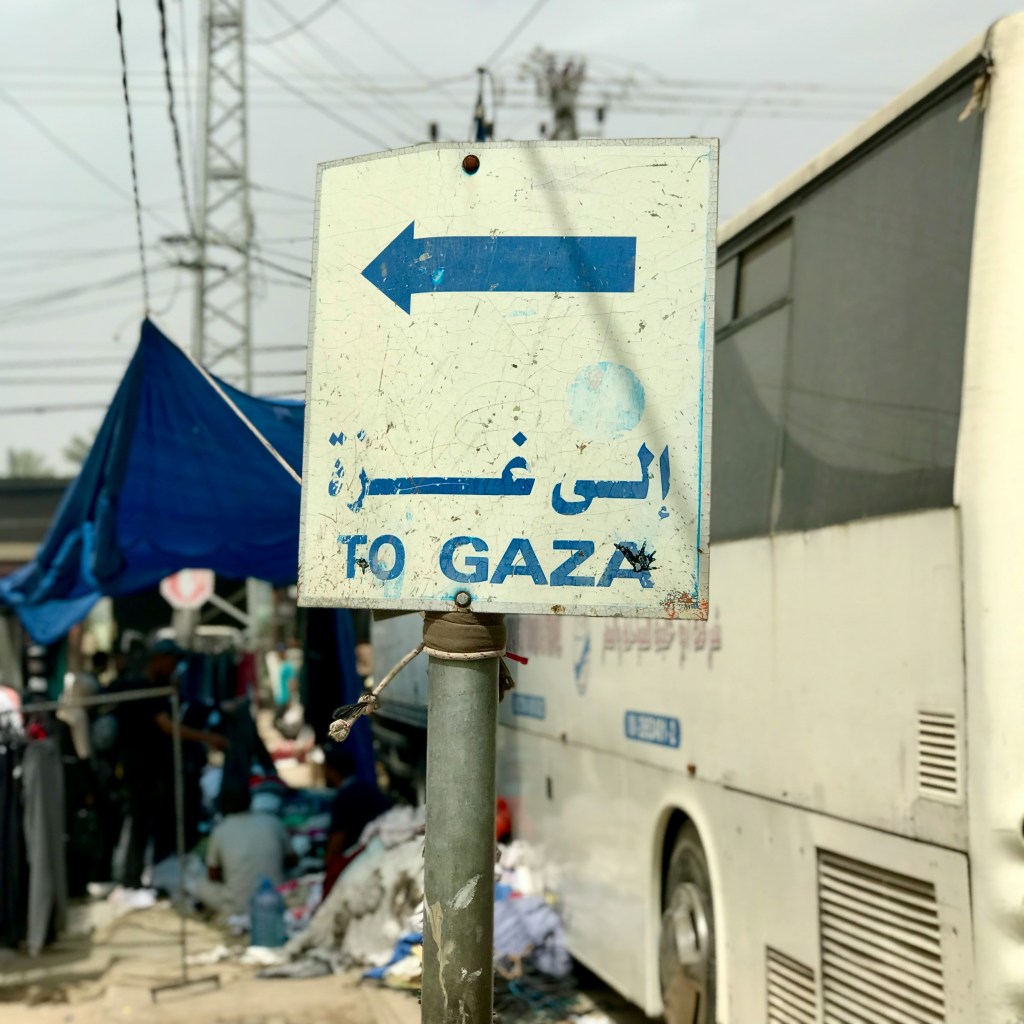 A bent road sign displaying the words To Gaza. Scenes of  displacement fill the background: a coach, its human and material contents spilling out onto the bare earth.
