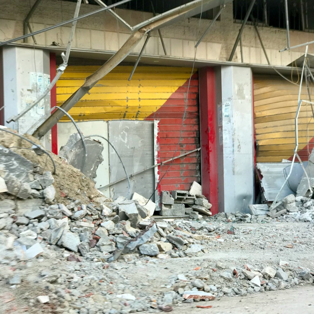 A bombed building in Gaza, red and yellow shutters to a shop next to rubble.