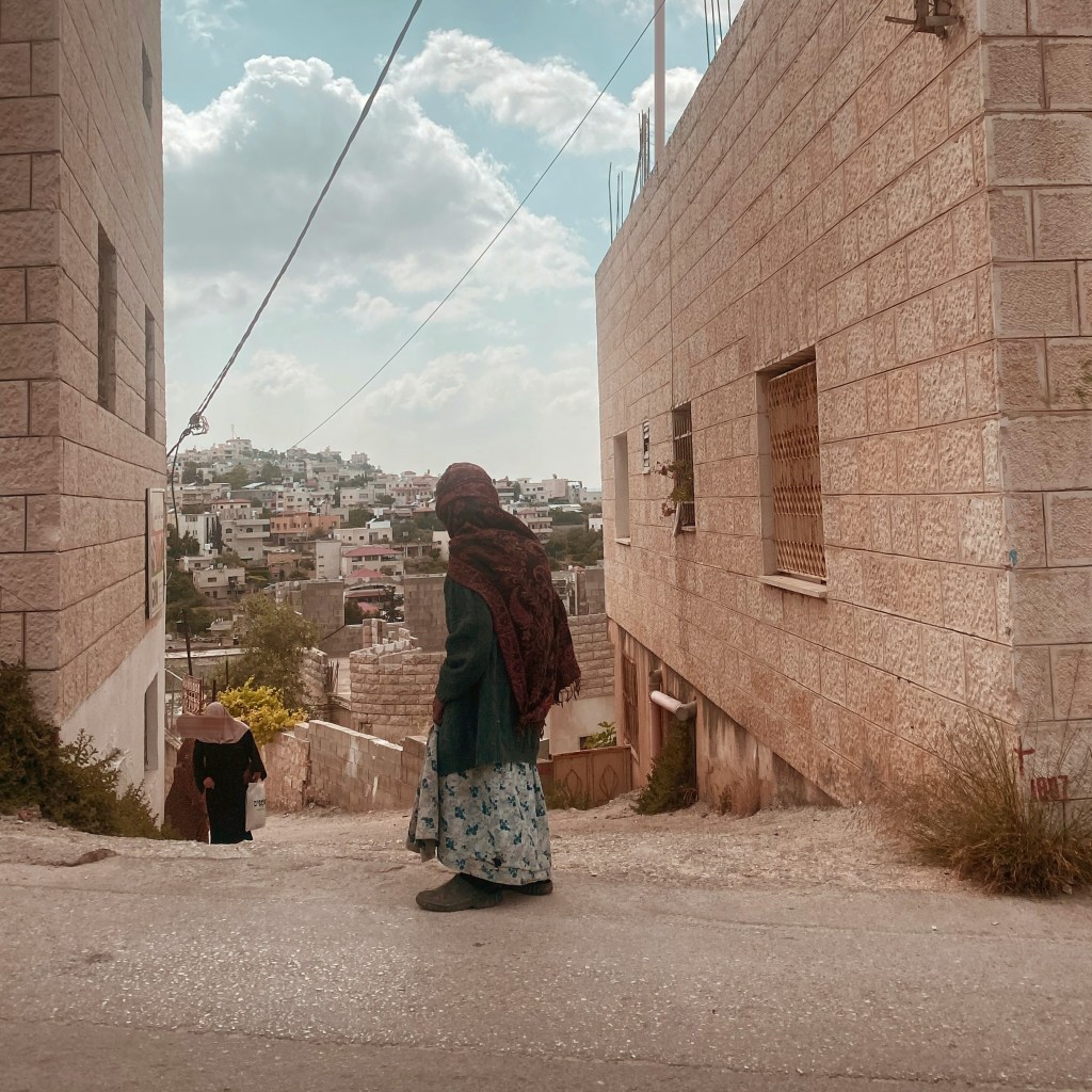 An elderly woman looks out at the view between two stone houses in Palestine.