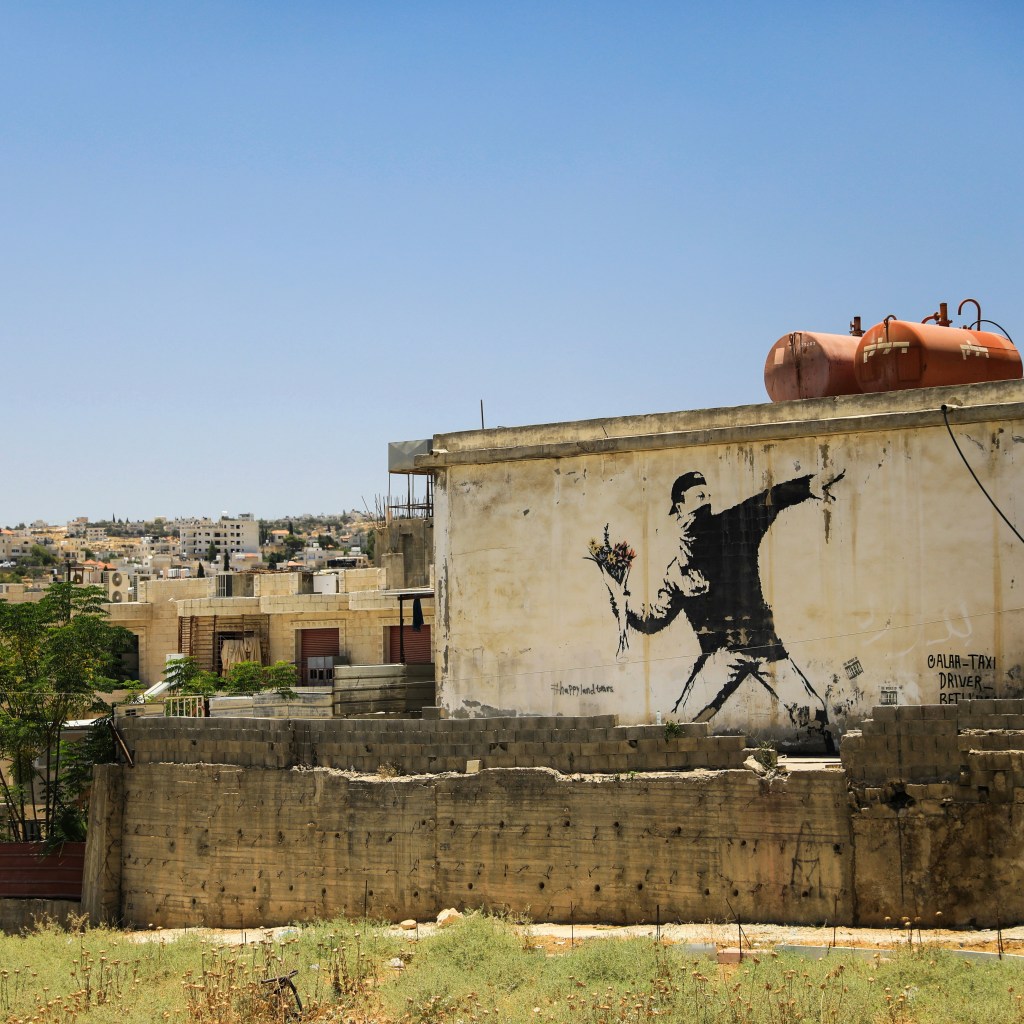A Banksy mural of a masked man throwing a bunch of flowers on the side of a flat-roofed building.