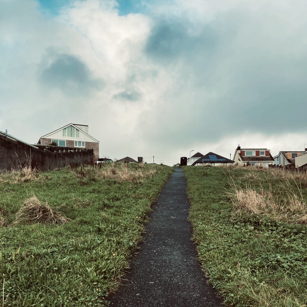 Looking uphill towards an estate of houses, the view split by a tarmacked path with grass on either side.