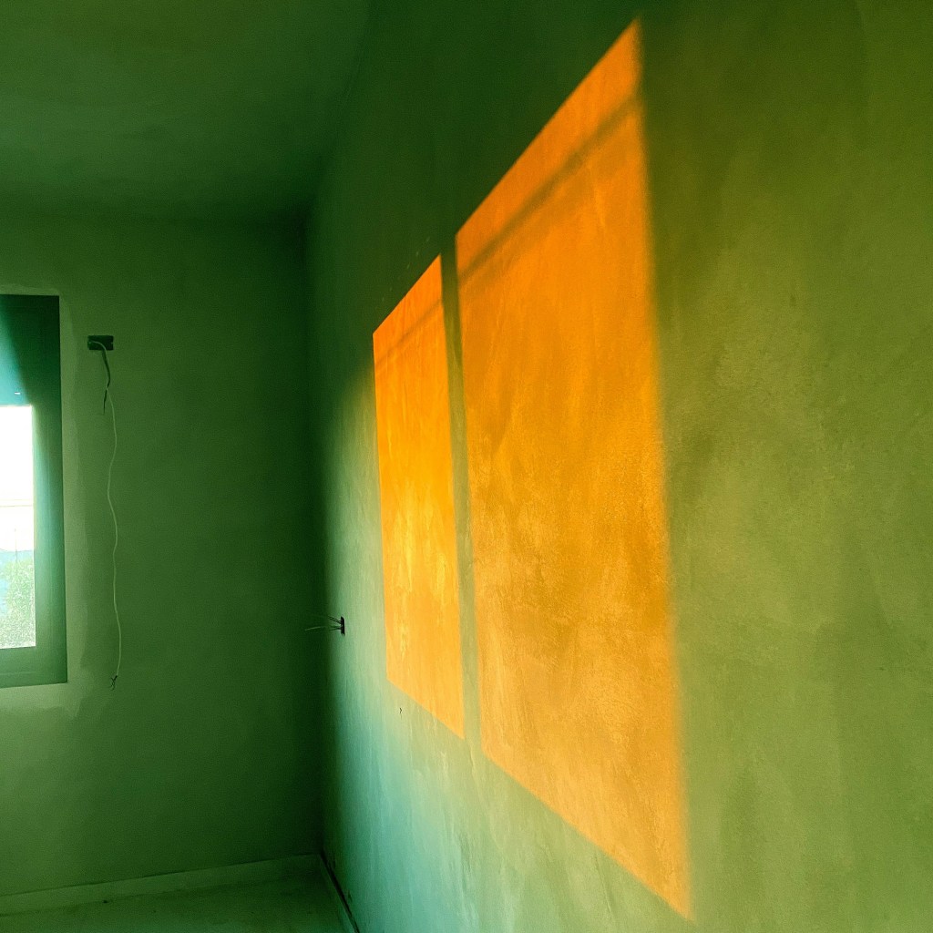 The orange outline of a window in shadow blazes on the green wall of an empty room